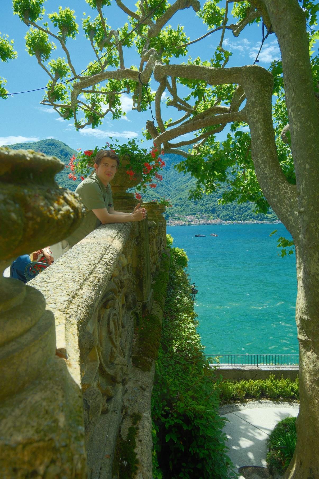 Brendan leaning on a stone balustrade beneath a tree in the gardens of Villa del Balbianello, the lake behind him