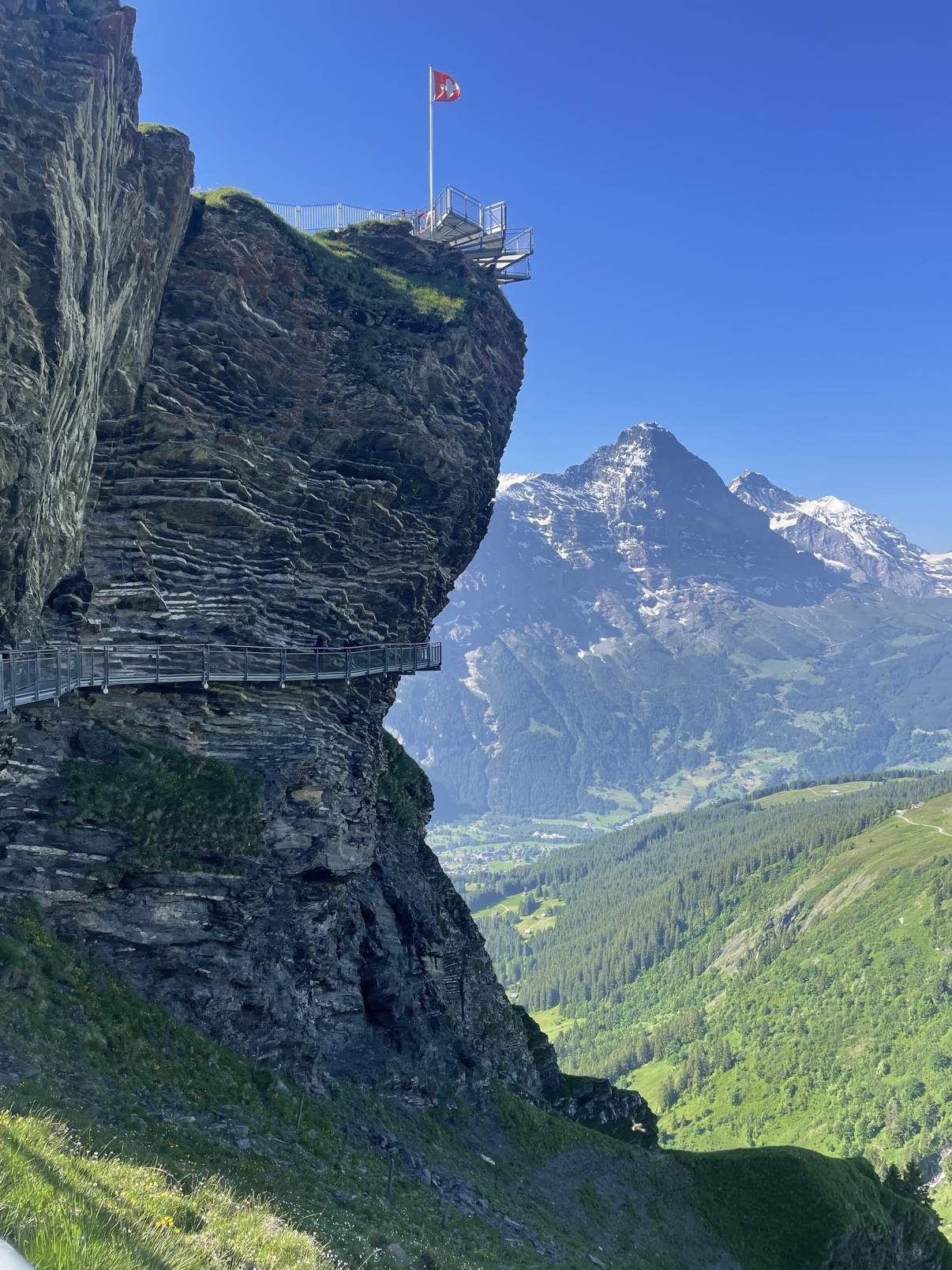 The metal cliff walkway at First, Grindelwald, with a Swiss flag flying and the valley far below